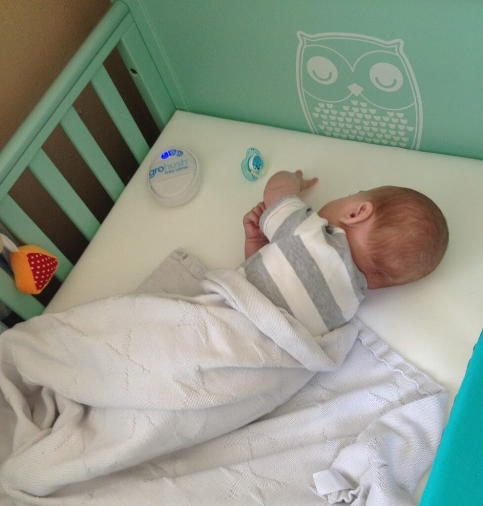A baby lies on their side in a mint green Cosatto cot bed, reaching towards a dummy. The baby is partially covered with a light blanket, and there is a small device and an owl decal on the cot wall.