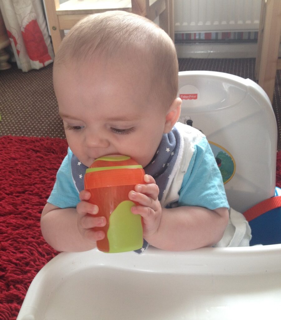 A baby in a highchair holds a green and orange beaker with both hands, bringing the beaker to their mouth—a perfect scene for weaning and introducing baby essentials. The baby wears a blue shirt and a dark bib with white stars.
