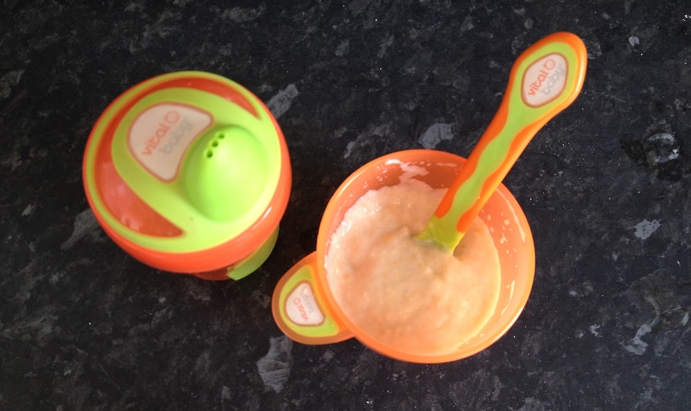 An orange and green vital baby sippy cup and a matching bowl with a spoon filled with baby food—a must-have for weaning—are placed on a dark worktop.