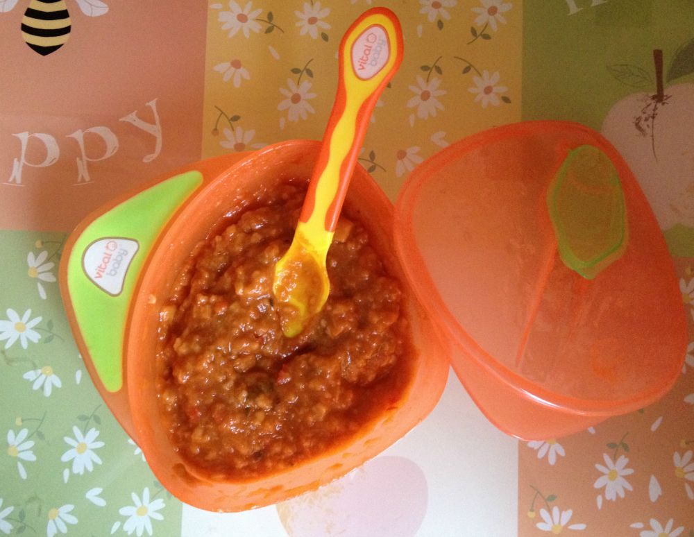 An orange bowl filled with mashed baby food, a colourful baby spoon inside—perfect for weaning. The bowl’s lid rests nearby on a floral-patterned surface with the word "Happy," capturing those baby essentials in style.