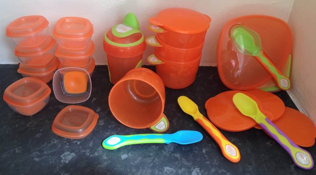 A collection of orange plastic food containers, bowls, and lids in various sizes, plus four colourful spoons—perfect for weaning or storing baby essentials—arranged on a dark worktop against a white wall.