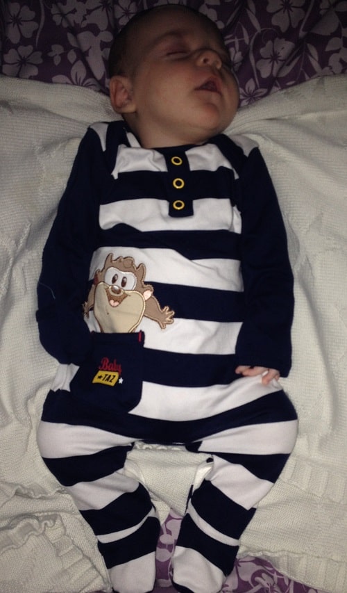 A baby sleeps on a blanket, wearing a striped navy and white outfit with a cartoon animal design and yellow buttons—an adorable scene that captures the peacefulness of baby sleep against a purple and white floral background.