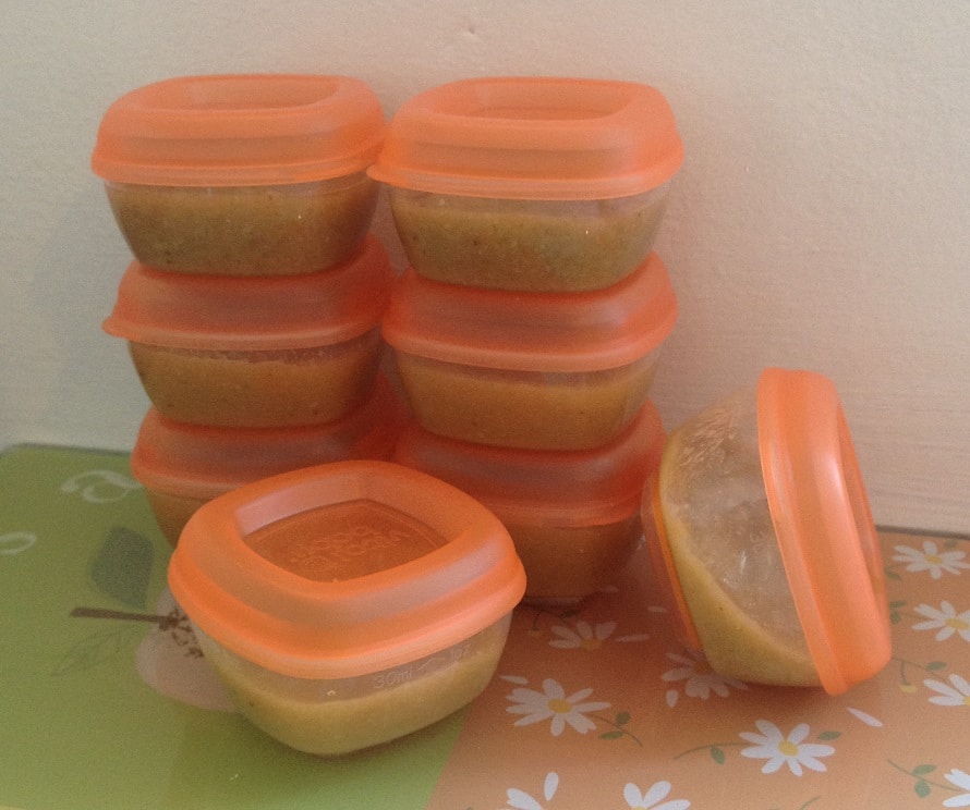 Eight small, orange-lidded plastic containers filled with food—perhaps Pasta Bake with Ricotta and Spinach—are stacked in two columns, with one container lying on its side. They rest on a table adorned with a floral and apple pattern.