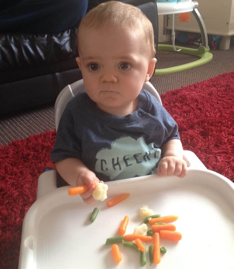 A baby sitting in a highchair looks up with a serious expression, holding a piece of cauliflower. Mixed vegetables like carrots, green beans, and cauliflower—and even hints of spinach—are on the tray in front of them.
