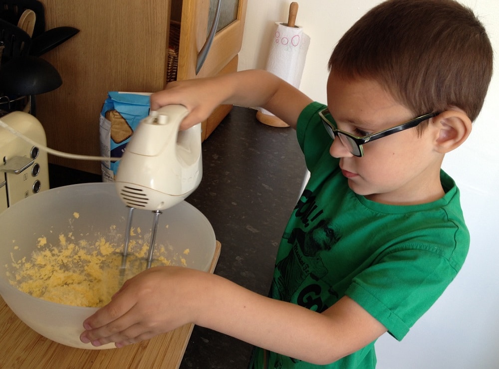 A young child wearing glasses and a green shirt uses a hand mixer to blend ingredients in a large bowl on a kitchen worktop while trying an easy buns recipe.
