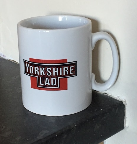 A white Yorkshire mug with a red and black "Yorkshire Lad" label sits on the edge of a dark worktop against a cream-coloured wall.