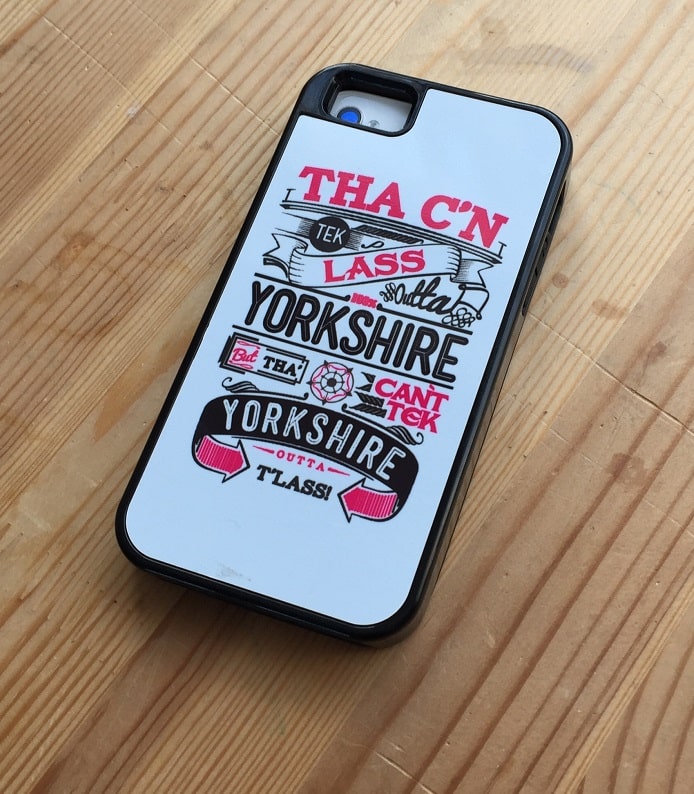 A smartphone in a black and white case featuring playful Yorkshire sayings rests on a wooden surface. The case displays colourful, varied fonts with phrases like "THA C'N TEK LASS OUTTA YORKSHIRE BUT THA CAN'T TEK YORKSHIRE OUTTA LASS.