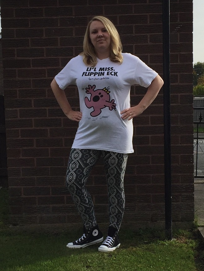 A woman stands outdoors in front of a brick wall, wearing a white "Lil Miss Flippin Eck" T-shirt—a classic Yorkshire phrase—patterned leggings, and black Converse trainers, with her hands on her hips and looking confidently at the camera.