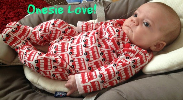 A baby lies on a cushioned seat wearing a red and white babygrow with a bus pattern. The words "Babygrow Love!" are written in green text, capturing a sweet moment in the Story Of Three Babies.