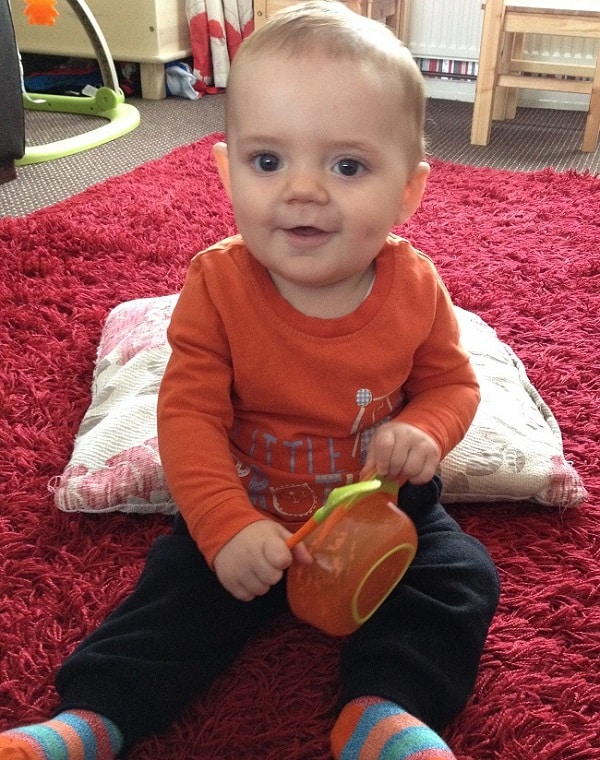 A baby wearing an orange shirt and black trousers sits on a red rug, holding an orange bowl and spoon—perfect for sharing baby weaning moments or seeking new weaning tips among colourful furniture and cosy cushions.