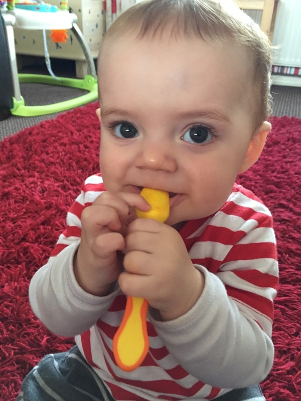 A baby in a red and white striped shirt sits on a red carpet, holding and chewing on a yellow and orange spoon—showcasing one of those adorable baby weaning moments with wide-eyed curiosity.