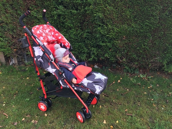 A baby wearing a hat and jacket sits in a black and red Cosatto Supa pushchair with star patterns, positioned on grass beside a tall, green hedge.