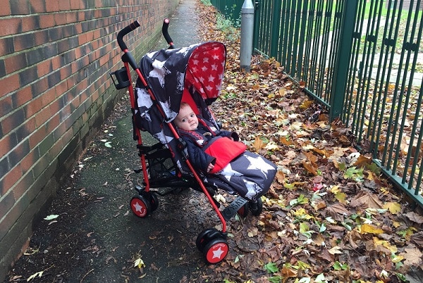 A baby sits in a Cosatto Supa Hipstar pushchair with a red and black design, along a path lined with a brick wall and metal fence, surrounded by fallen autumn leaves.