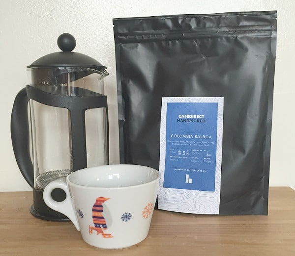 A cafetière, a white mug with a dog and snowflakes, and a black bag of Colombia Balboa coffee with a blue label are arranged on a wooden surface—perfect inspiration for your Christmas Gift Guide or Gifts for Men.