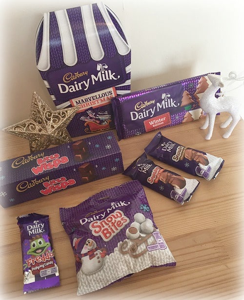 A selection of Cadbury Dairy Milk chocolate treats, including bars, bags, and boxes, arranged on a wooden surface with festive decorations—perfect for younger boys' Christmas gift ideas.