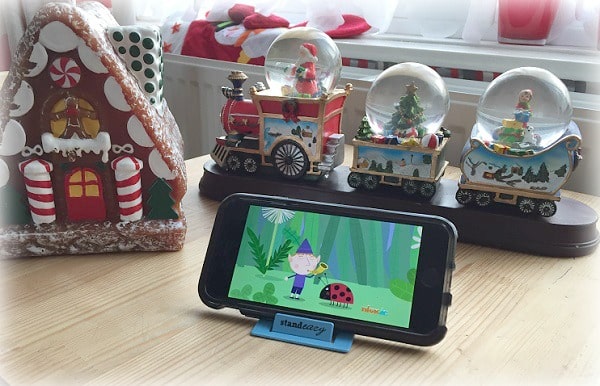 A smartphone on a stand plays a colourful cartoon featuring a ladybird. Beside it, Christmas Gift Guide favourites—a gingerbread house decoration and a festive train with three snow globes—sit on a wooden table.