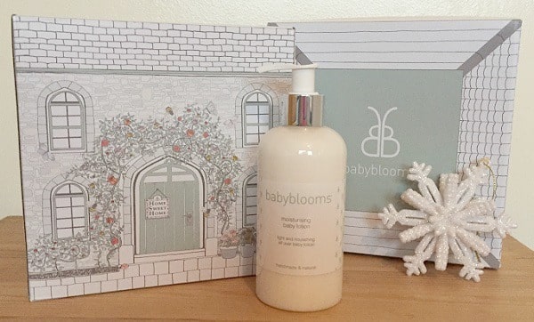 A bottle of Babyblooms moisturising baby lotion is displayed in front of a decorative box with a house illustration, next to a white snowflake ornament—perfect for younger boys’ Christmas gift ideas—on a wooden surface.