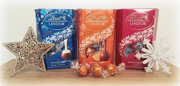 Three boxes of Lindt Lindor chocolates (blue, orange, and red) are displayed on a wooden surface, flanked by a gold star and a white snowflake—perfect for Christmas Gifts or to feature in your Christmas Gift Guide. Unwrapped chocolates rest in front.