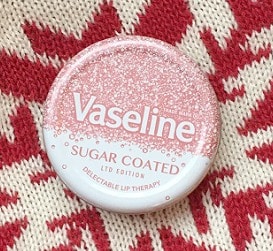 A round tin of Vaseline Sugar Coated Limited Edition lip therapy with a pink and white sparkly design, placed on red and cream knitted fabric—perfect for any Christmas Gift Guide or as a festive treat in your collection.