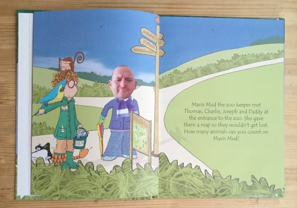 A children’s storybook illustration shows three characters by a zoo entrance sign. One sports an adult human's face edited onto their body—perfect for quirky Christmas Gifts for Men. There’s a map, signpost, grass, and path in the background.
