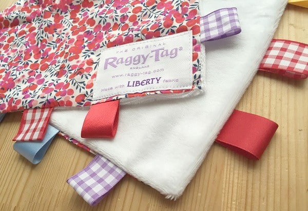 A soft, white baby comforter with a colourful floral pattern and ribbon tags, perfect for younger boys’ Christmas gift ideas. The label reads "Raggy-Tag" and "Made with Liberty Fabric," shown lying on a wooden surface.