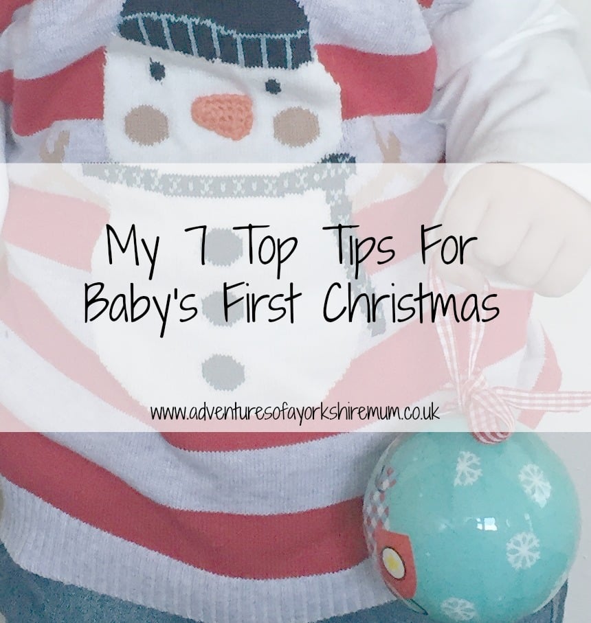 A baby wearing a festive jumper with a snowman design holds a blue Christmas bauble. Text overlay reads, "My 7 Top Baby Christmas Tips for Baby's First Christmas," with a website link below.