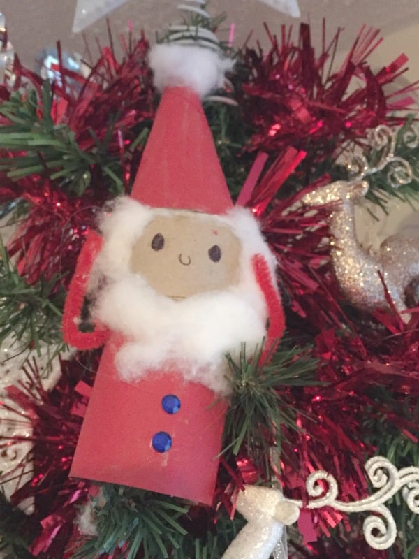 A homemade Father Christmas ornament made from a red-painted cardboard tube, cotton beard, and red hat hangs on a Christmas tree adorned with holiday decorations like red tinsel and a silver reindeer, capturing the love of Christmas.