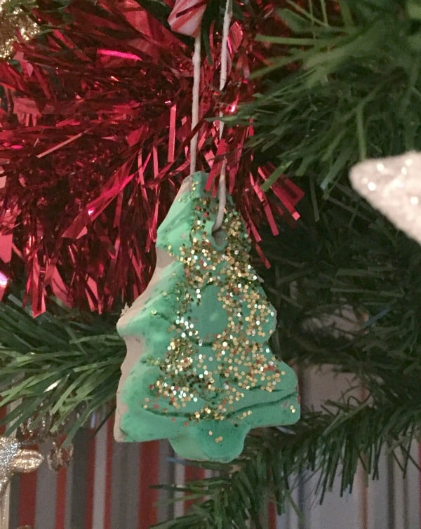 A Christmas tree-shaped ornament decorated with green icing and gold glitter hangs on a Christmas tree, surrounded by festive Christmas decorations, red tinsel, and lush green branches in the background.