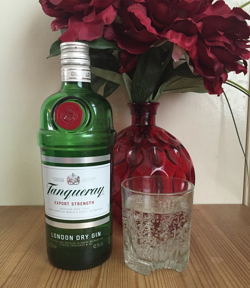 A green bottle of Tanqueray London Dry Gin stands next to a glass of clear liquid and a red vase with large red flowers on a wooden surface—an elegant addition to any Christmas Gift Guide.
