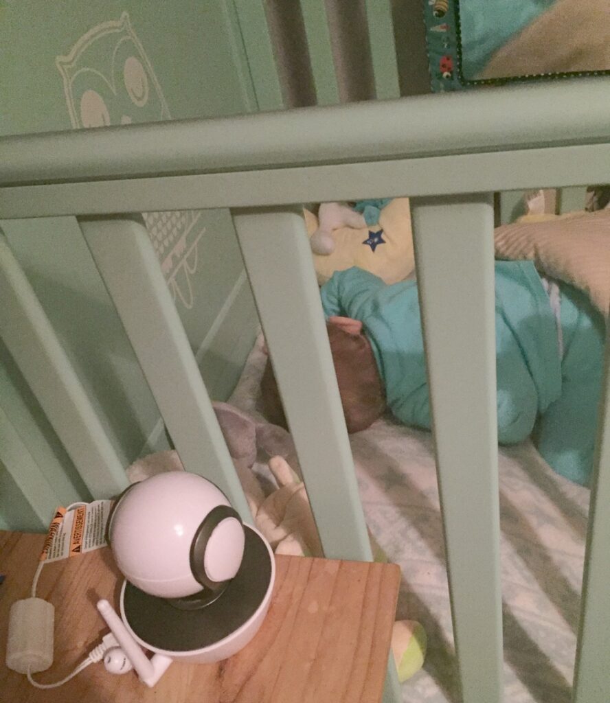 A baby sleeps on their side in a cot with pale green bars, surrounded by soft blankets and toys. A Motorola MBP853 baby monitor rests on a wooden table next to the cot. An owl design is visible on the wall.