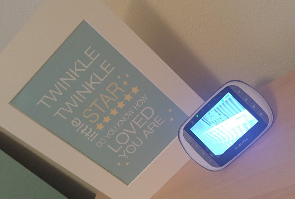 A framed print with the words "Twinkle Twinkle Little Star, Do You Know How Loved You Are" sits next to a Motorola MBP853 Connect baby monitor displaying a cot on a light wooden surface.