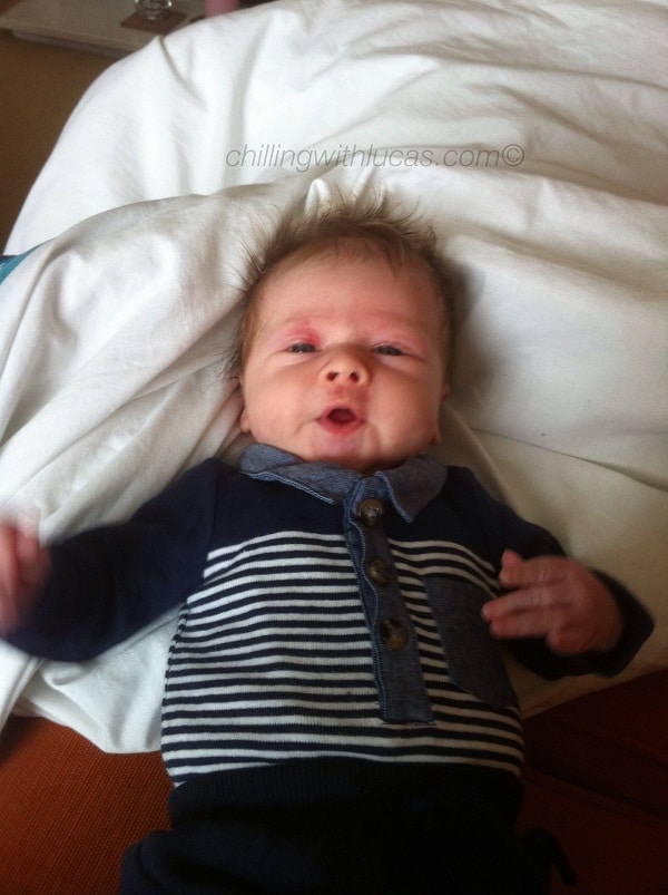 A baby with spiky hair lies on a bed dressed in a navy blue outfit with horizontal white stripes and buttons, looking up with mouth slightly open—a sweet reminder of the joys that follow good pregnancy advice.