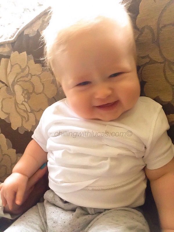 A smiling baby with blond hair sits on a patterned sofa, wearing a white babygrow and grey trousers. Bathed in natural sunlight, this joyful scene reminds us of the special moments to look forward to—one of many things to know when pregnant.