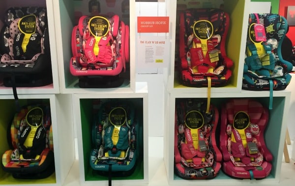 Six colourful Cosatto child car seats with playful patterns are displayed in individual cubby shelves; three seats are on the top row, three on the bottom. A sign with text is positioned in the middle of the Cosatto display.