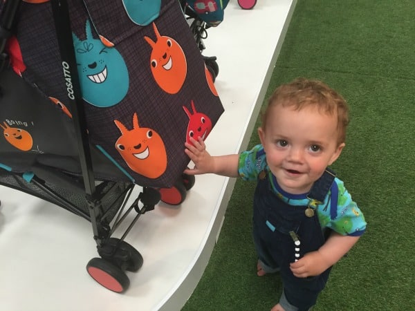A smiling toddler wearing blue dungarees stands next to a Cosatto pushchair decorated with colourful, cartoonish faces. The scene is indoors, set atop green artificial grass flooring.