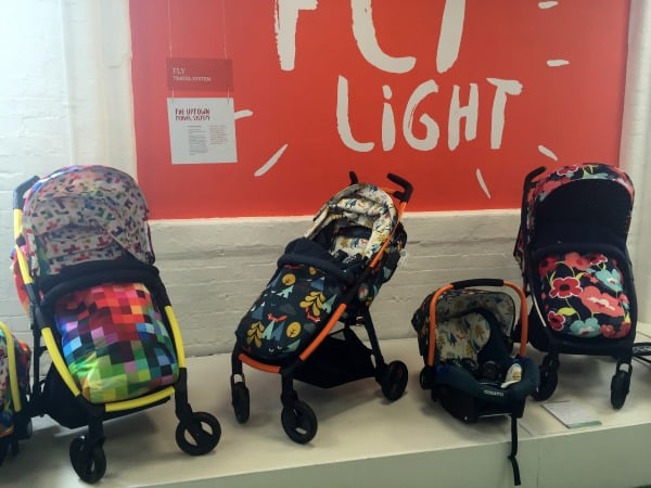 Four colourful Cosatto pushchairs, featuring pixel art, cartoons, and floral patterns, are displayed in a row on a white platform in front of a red wall with “FLY LIGHT” written in white.