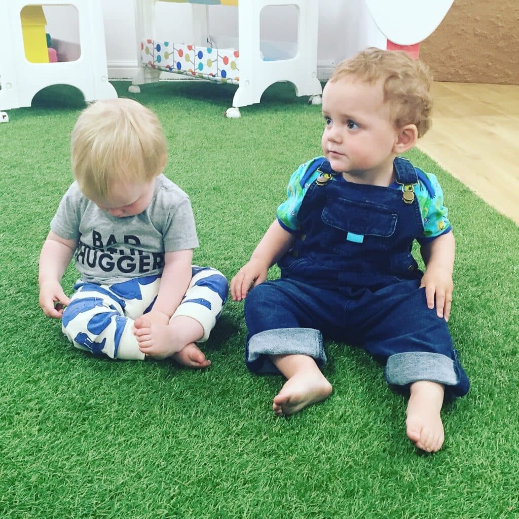 Two toddlers sit barefoot on artificial green grass. One, in a grey "BAD HUGGER" shirt and white patterned trousers, looks down; the other, in a blue shirt and denim dungarees, looks to the side next to their colourful Cosatto pushchair.