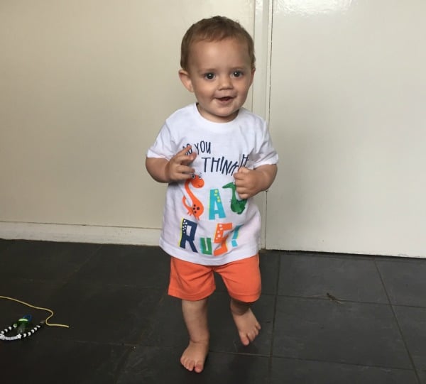 A smiling toddler with light brown hair stands on a tiled floor, wearing orange shorts and a white WTBW T-shirt with colourful letters. Perfect for summer fashion, the child appears mid-step, with a toy visible on the floor nearby.