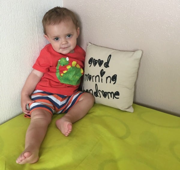 A young child in a red shirt and striped shorts, perfect for Summer Fashion, sits on a green surface next to a cushion that reads "good morning handsome." The child is smiling slightly and leaning against the wall.