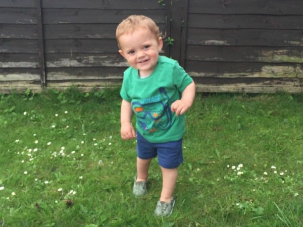 A young child with short light brown hair, dressed in summer fashion—a green T-shirt, blue shorts, and grey shoes from Mothercare—stands smiling on green grass in front of a dark wooden fence.
