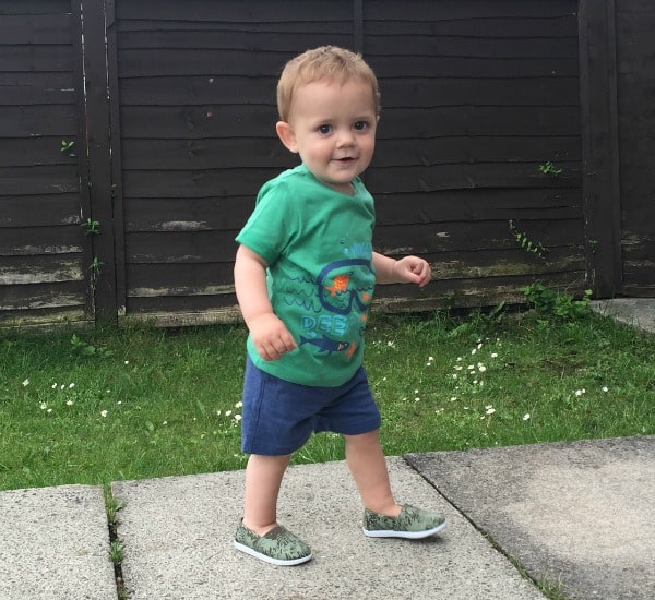 A young toddler with light brown hair, dressed in summer fashion from Mothercare—a green T-shirt, blue shorts, and green slip-on shoes—is walking outdoors on a stone path beside a grassy lawn and dark wooden fence.