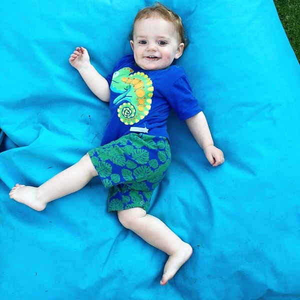 A smiling toddler lies on a bright blue blanket outdoors, dressed in the Rockin' Baby Out Of Africa Romper featuring a colourful chameleon graphic and green shorts with a leaf pattern.