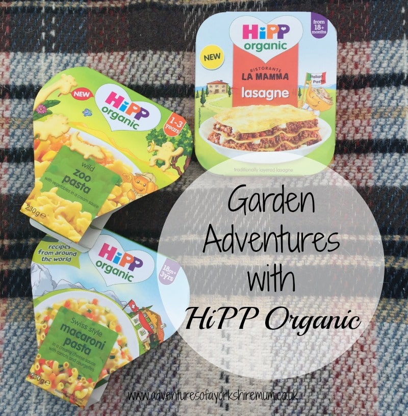 Three HiPP Organic baby food packs (lasagne, zoo macaroni pasta, and Swiss-style macaroni cheese) arranged on a checked blanket with the text "Garden Adventures with HiPP Organic" in the centre, perfect for little ones who love organic gardening fun.