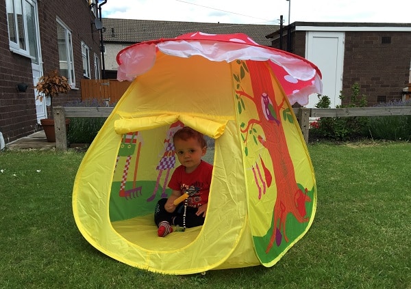 A young child sits inside a colourful, mushroom-shaped play tent on a grassy lawn beside a brick house. The tent, perfect for Garden Adventures, is decorated with animal illustrations and has a yellow entrance flap.