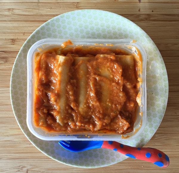 A plastic container filled with three cannelloni covered in tomato sauce sits on a plate with a blue and red spotty spoon, all placed on a wooden surface—a perfect meal for little ones enjoying their HiPP Organic garden adventures.