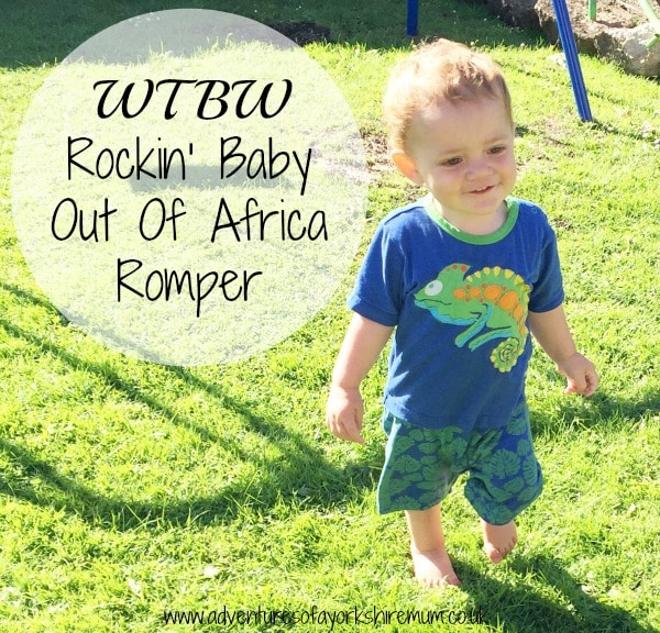 A toddler in a blue Out Of Africa romper with a green chameleon design stands on sunlit grass. Text reads: "WTBW Rockin' Baby Out Of Africa Romper." A playground swing is seen in the background.