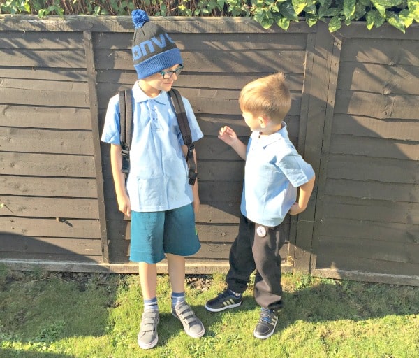 Two young boys in school uniforms stand outside by a wooden fence, looking P.E. ready. One sports a hat, glasses, rucksack, and shorts from Get The Label; the other wears black trousers. They appear to be interacting with each other.