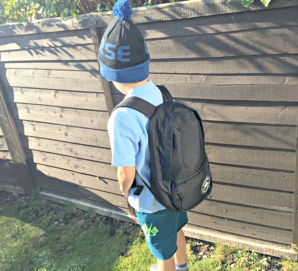 A child wearing a blue shirt, blue shorts, and a blue and black beanie stands outdoors near a wooden fence with a black rucksack, looking P.E. Ready. Kit your kids out for any adventure with Get The Label style.