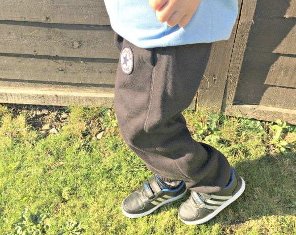 A child in dark tracksuit bottoms, a light blue top, and black trainers with white soles stands on grass near a wooden fence. The sun casts bright light across the P.E. Ready scene—perfect for active days with Get The Label style.