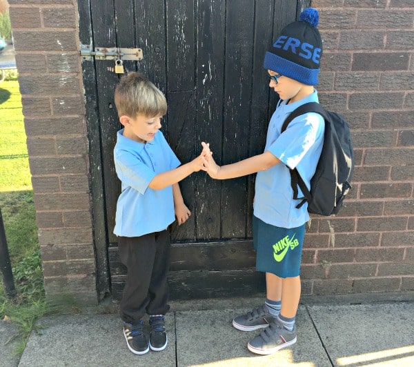 Two young boys in blue shirts stand facing each other outside, looking P.E. ready. One, in sunglasses, a beanie, and shorts with a rucksack from Get The Label; the other in long trousers. They touch hands by a dark wooden door and brick wall.
